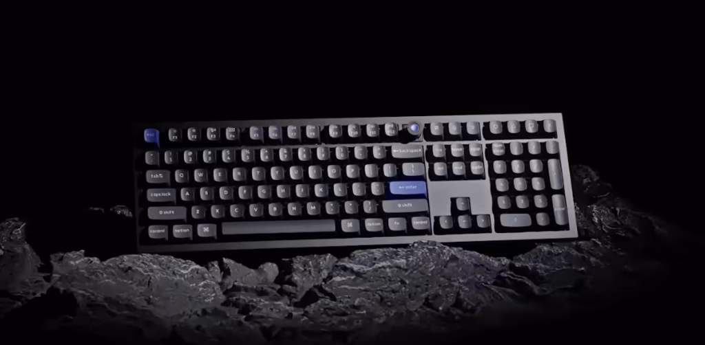 Keychron Q6 HE 8K, tastiera magnetica full-size per lavoro e gaming Mechanical keyboard with blue keycaps resting on a rocky surface in a dark setting.