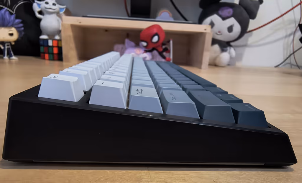 Low-angle side view of a two-tone mechanical keyboard with white and dark gray keys on a wooden desk; blurred toys in the background.