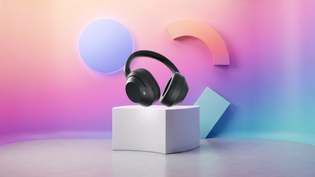Black over-ear headphones resting on a white cube pedestal against a pastel gradient backdrop with geometric shapes in the background, product shot